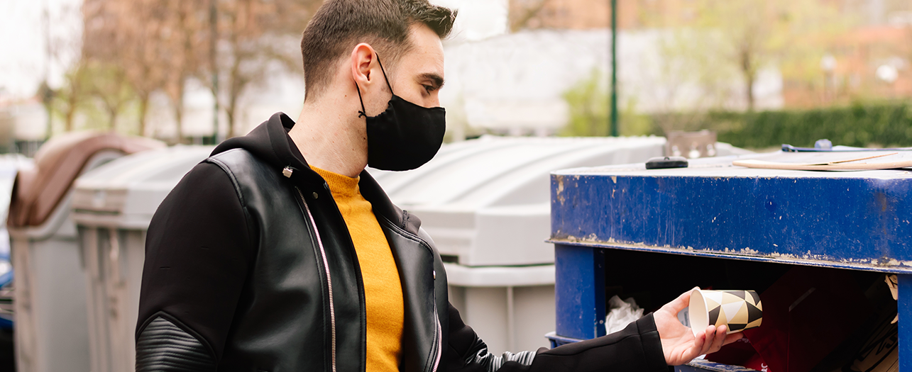 A man wearing a mask is throwing trash into an outdoor bin