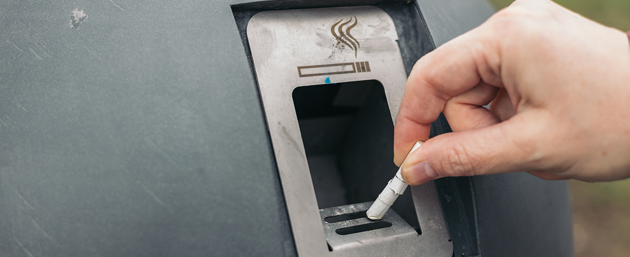 Hand disposing of a cigarette into an outdoor ashtray bin.